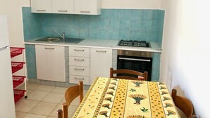 A compact kitchen with white cabinets, a gas stove, a sink, and a small oven. There is a dining table with chairs and a red basket near the sink. - Villa Arco 5, Palinuro, Cilento (Palinuro)
