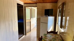 Quarto pequeno com cama, uma mesa com toalha de mesa florida, televisão sobre suporte e um frigorífico.