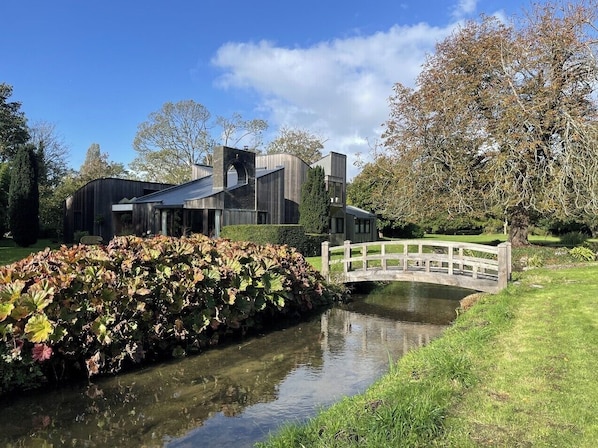A modern building with a large glass facade, a small bridge, and a canal with water plants. - Unique, Annexe sleeps 4 with Large Gardens & Tennis Court! (Chichester)
