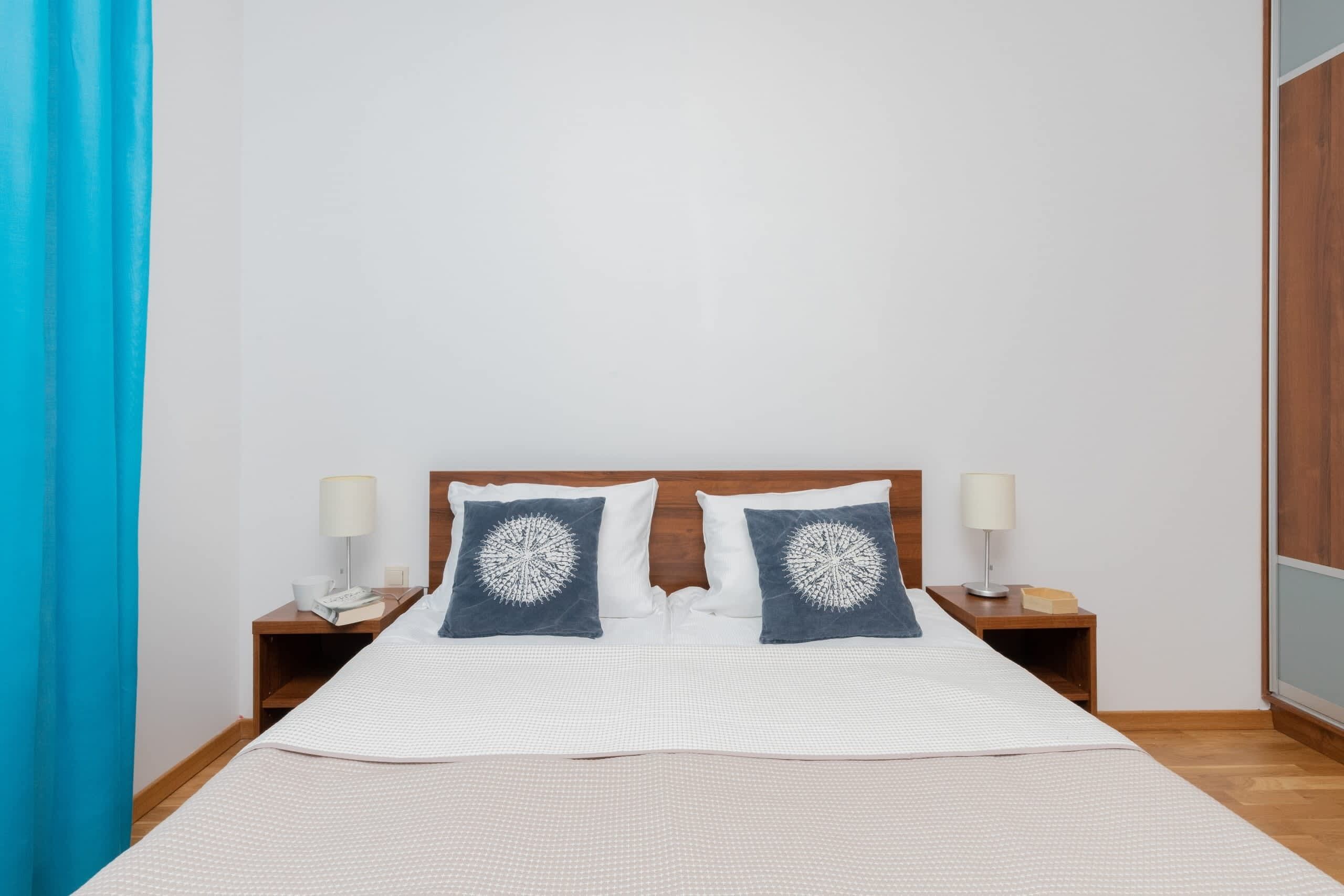 Een net opgemaakt bed met een wit-blauwe spreideken, twee bijpassende kussens, en twee nachtkastjes met lampen en een telefoon.