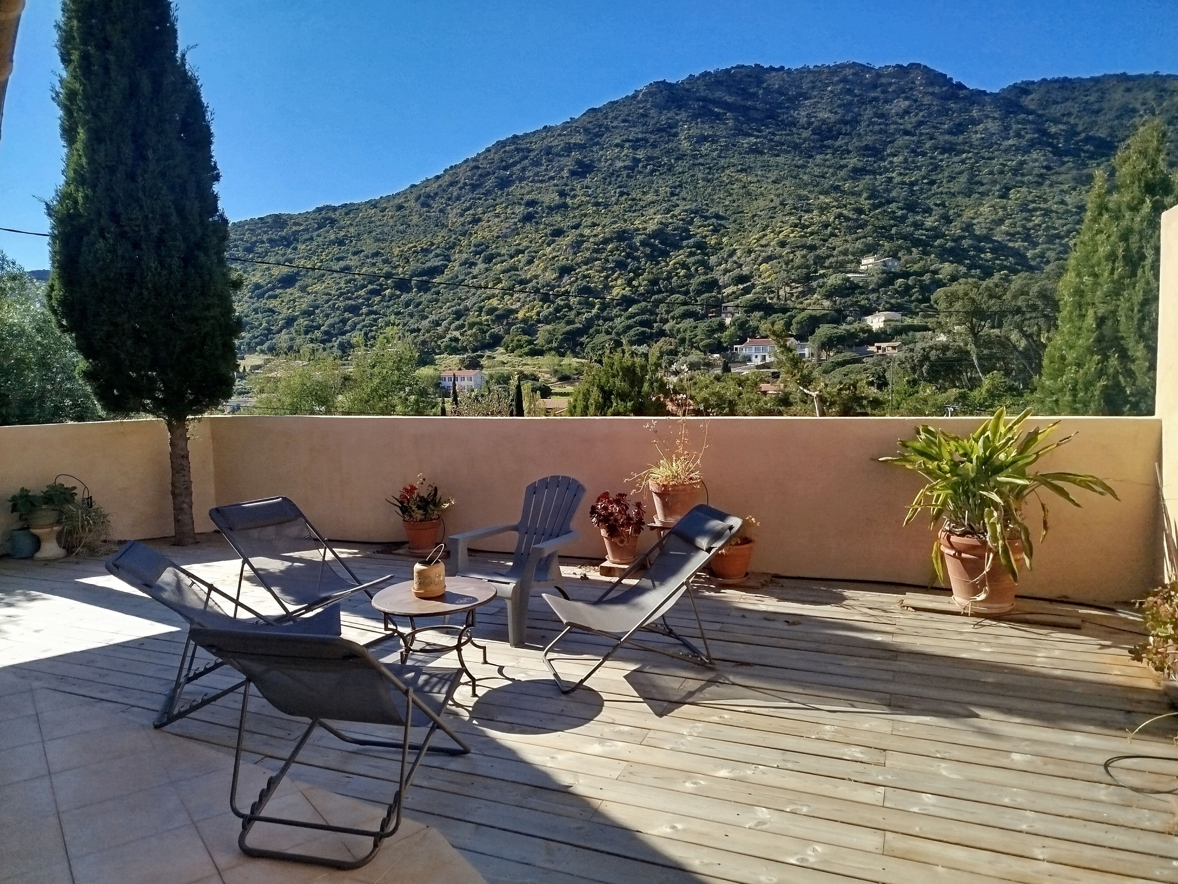 A wooden deck with lounge chairs and a table, surrounded by potted plants, overlooking a mountainous landscape.