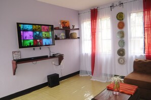 A living room with a flat-screen TV mounted on the wall, a wooden shelf with decorative items, a red and white curtain, and a brown sofa. - Hakuna Matata Cozy Apartment (Karatu)