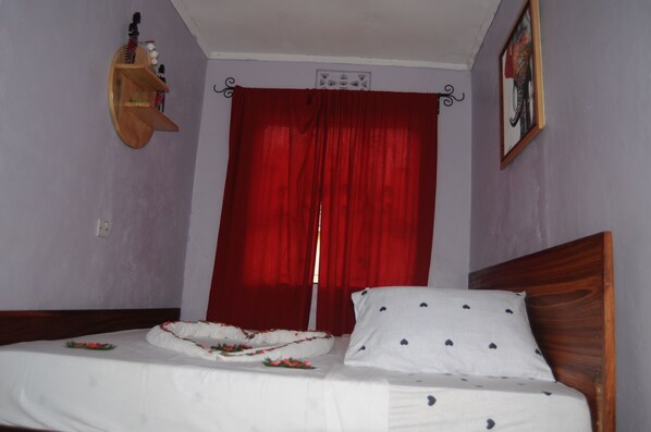 A bedroom with a wooden headboard, a bed with white bedding and a floral pillow, a red curtain, and a small shelf with decorative items. - Hakuna Matata Cozy Apartment (Karatu)