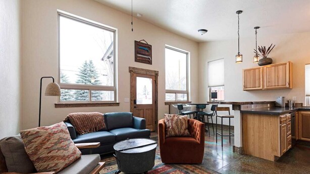A spacious living room with a sofa, armchair, and coffee table, a kitchen area with wooden cabinets, and large windows offering a view of trees. - Rustic Modern Home in East Missoula (Missoula)