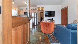 A modern living room with a blue sofa, a wooden door, a kitchen with cabinets and a refrigerator, and a television mounted on the wall. - Rustic Modern Home in East Missoula (Missoula)