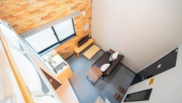 A compact room with a wooden wall, a sofa, a small table, a window, and a kitchenette area.