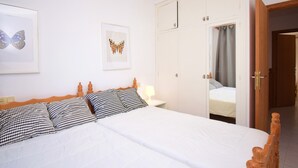Una cama con ropa de cama blanca, un cabecero de madera y dos almohadas a cuadros de galgo. Hay una mesita de noche con una lámpara y una obra de arte con una mariposa enmarcada en la pared. Se ve un armario con un espejo y una puerta.