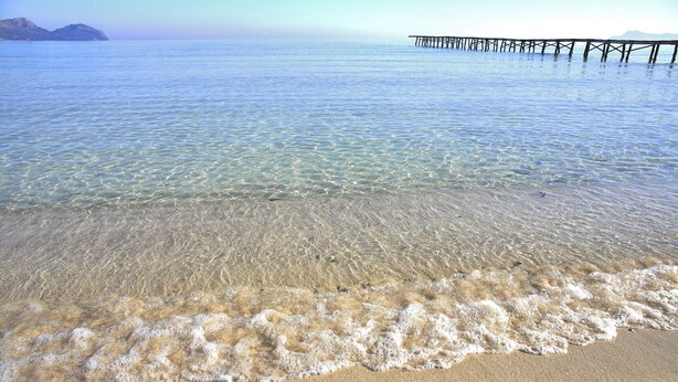 Una playa con aguas transparentes y poco profundas, una orilla arenosa y un muelle que se adentra en el agua.