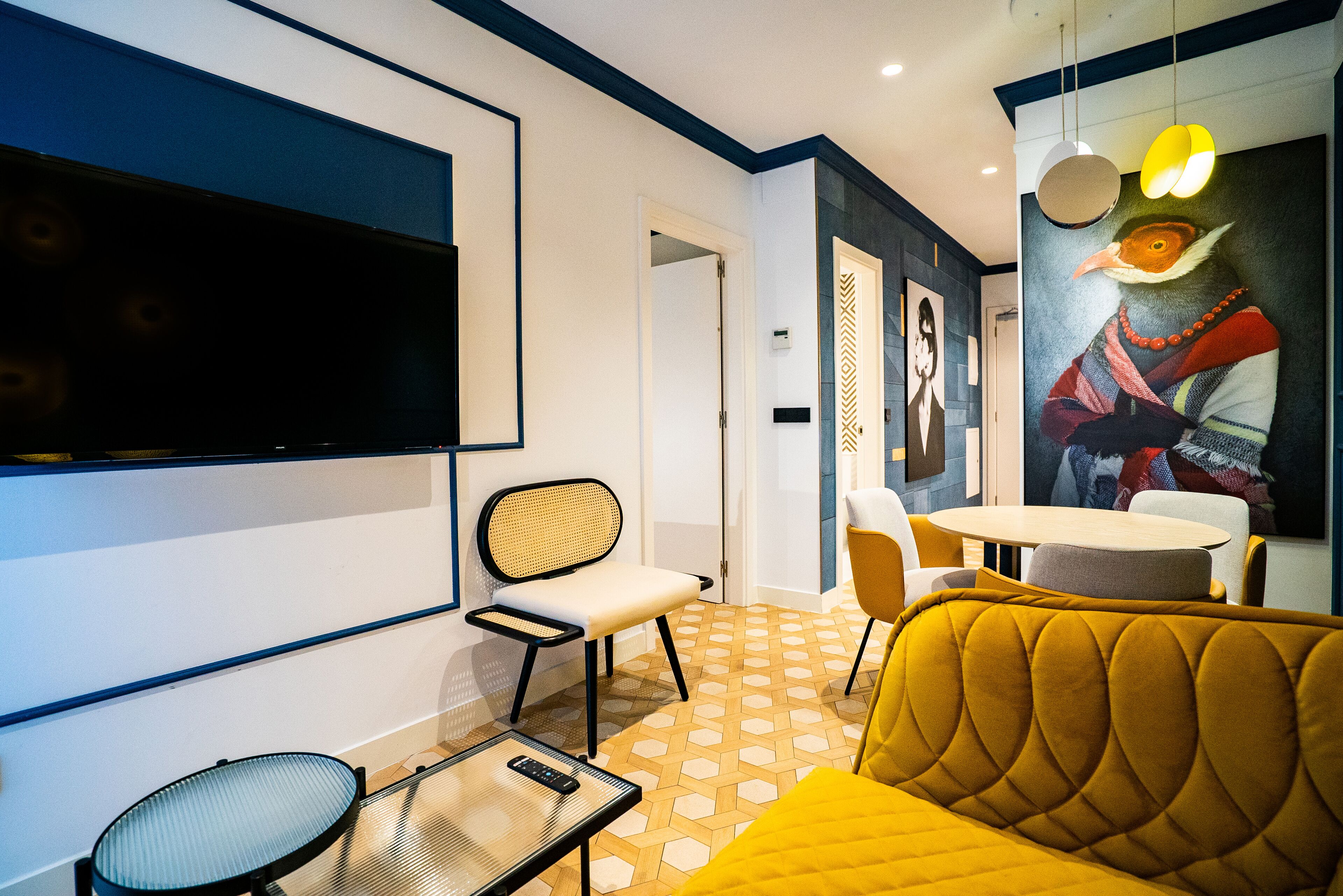 A modern living room with a flat-screen TV, a yellow sofa, a dining area with chairs, and a patterned floor.