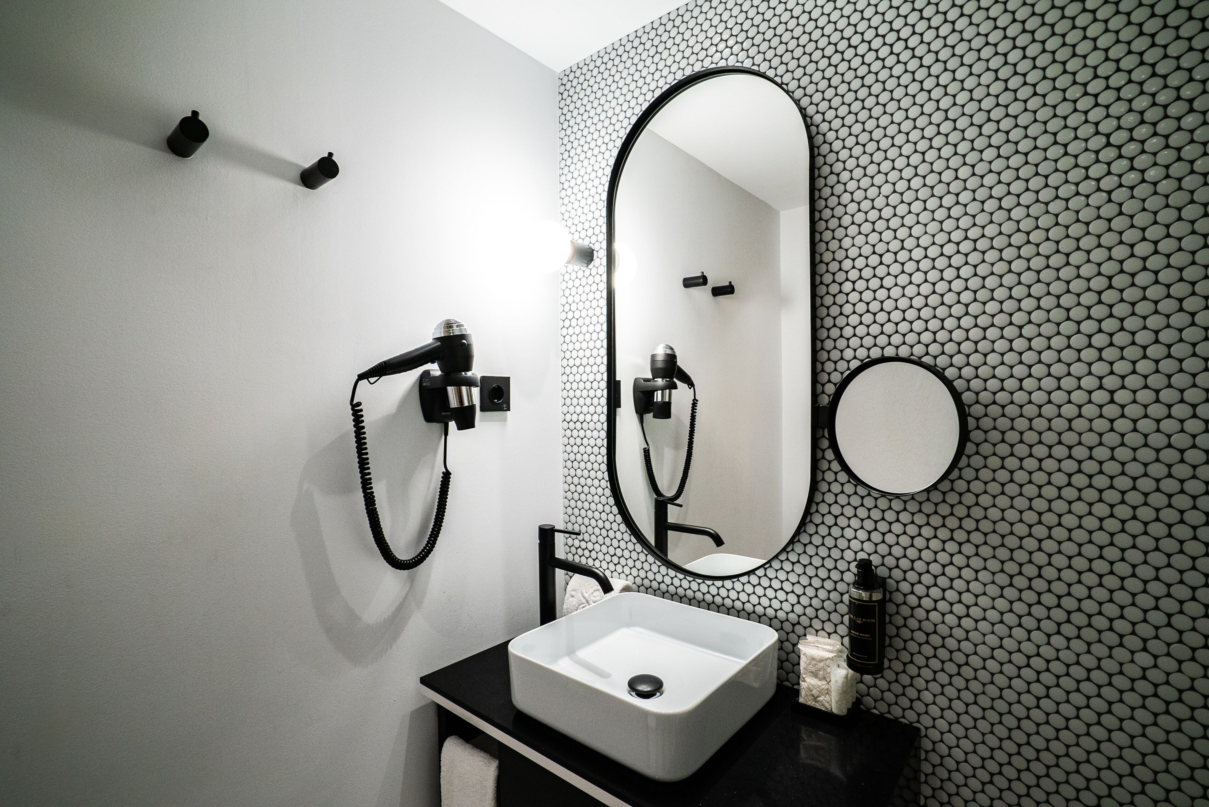 Un baño moderno con pared de azulejos hexagonales en blanco y negro, un lavamanos rectangular, un espejo ovalado grande y un cabezal de ducha de mano.
