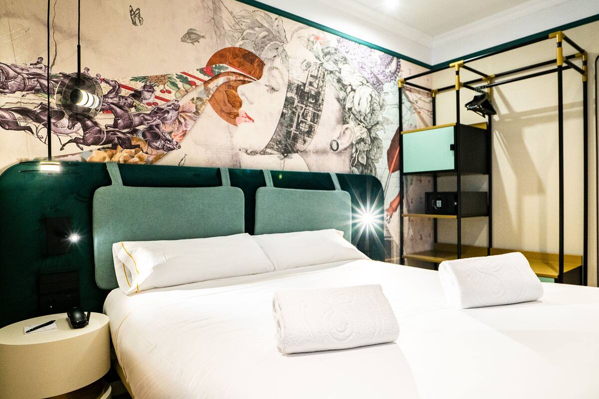 A hotel room with a large bed, a headboard with artwork, a bedside table with a lamp, and a small shelf.