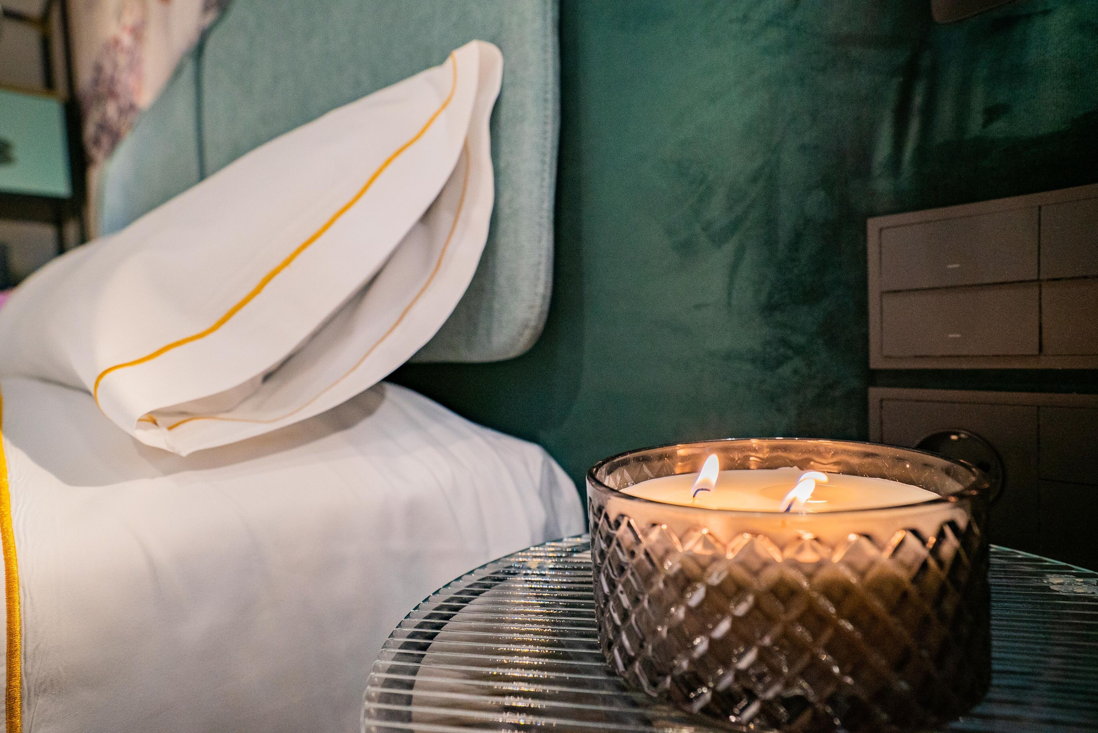 Una cama con ropa de cama blanca y un almohadón blanco con una franja amarilla, un candelabro de vidrio con una vela encendida, y una pared verde al fondo.