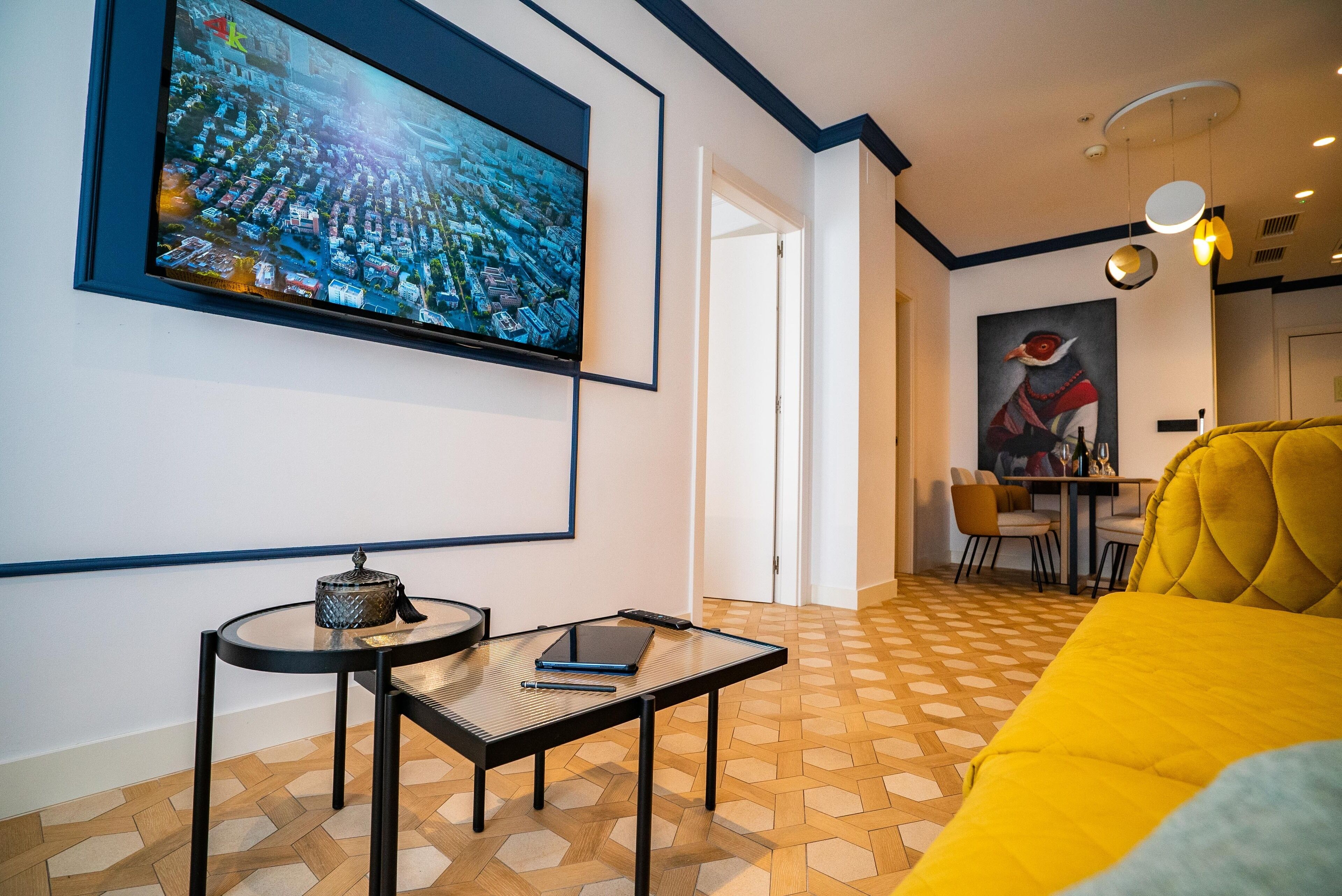 A modern living room with a large flat-screen TV mounted on the wall, a small round table with a laptop and a decorative item, a yellow sofa, and a dining area with a table and chairs.