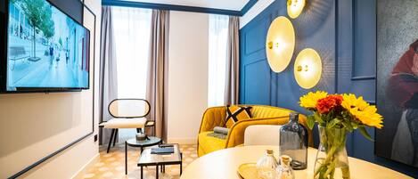 A modern hotel room with a large TV, a sofa, a dining table with a vase of flowers, and a wall-mounted light fixture.