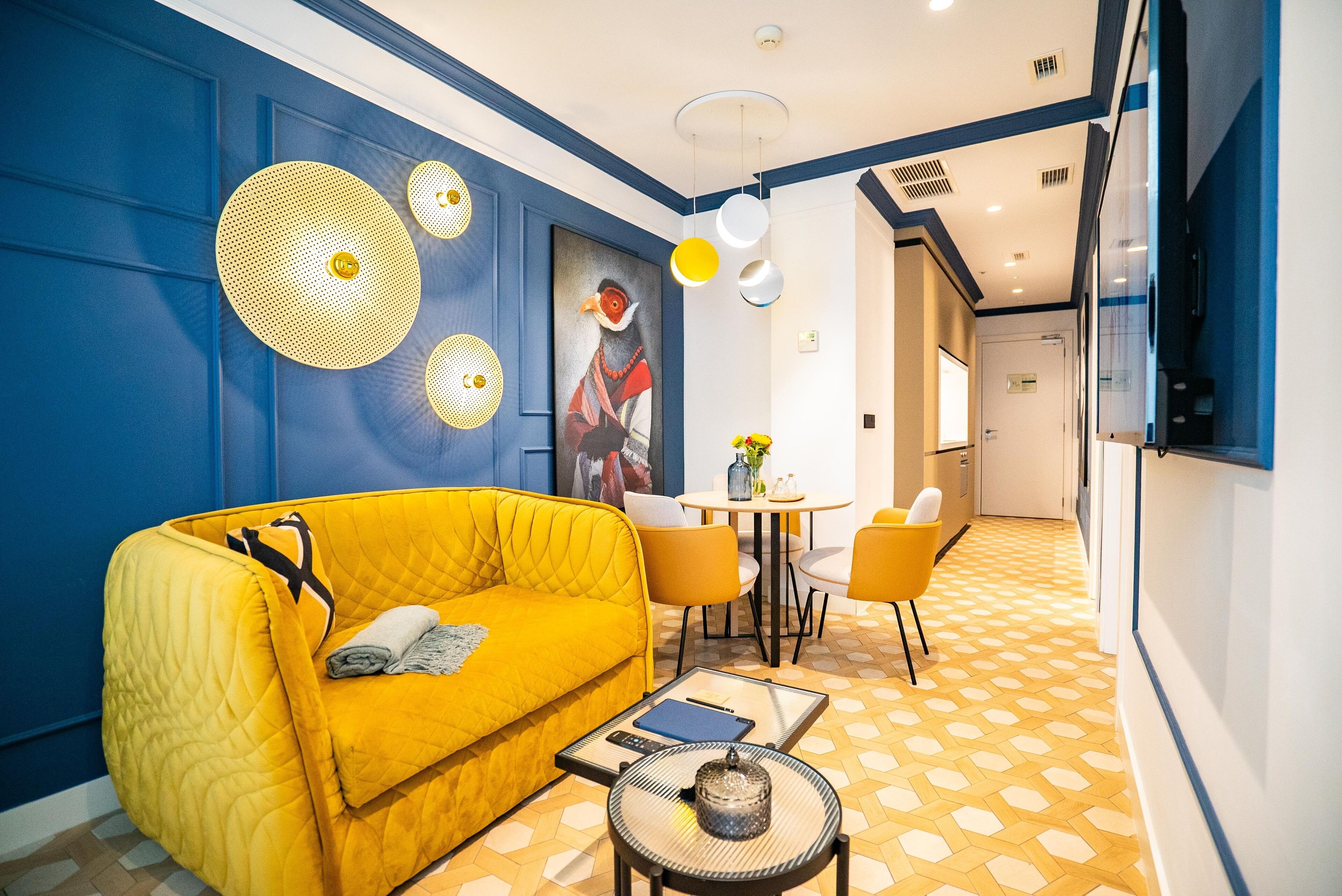 A modern hotel room with a yellow sofa, a round coffee table, and a dining area with a small table and chairs.