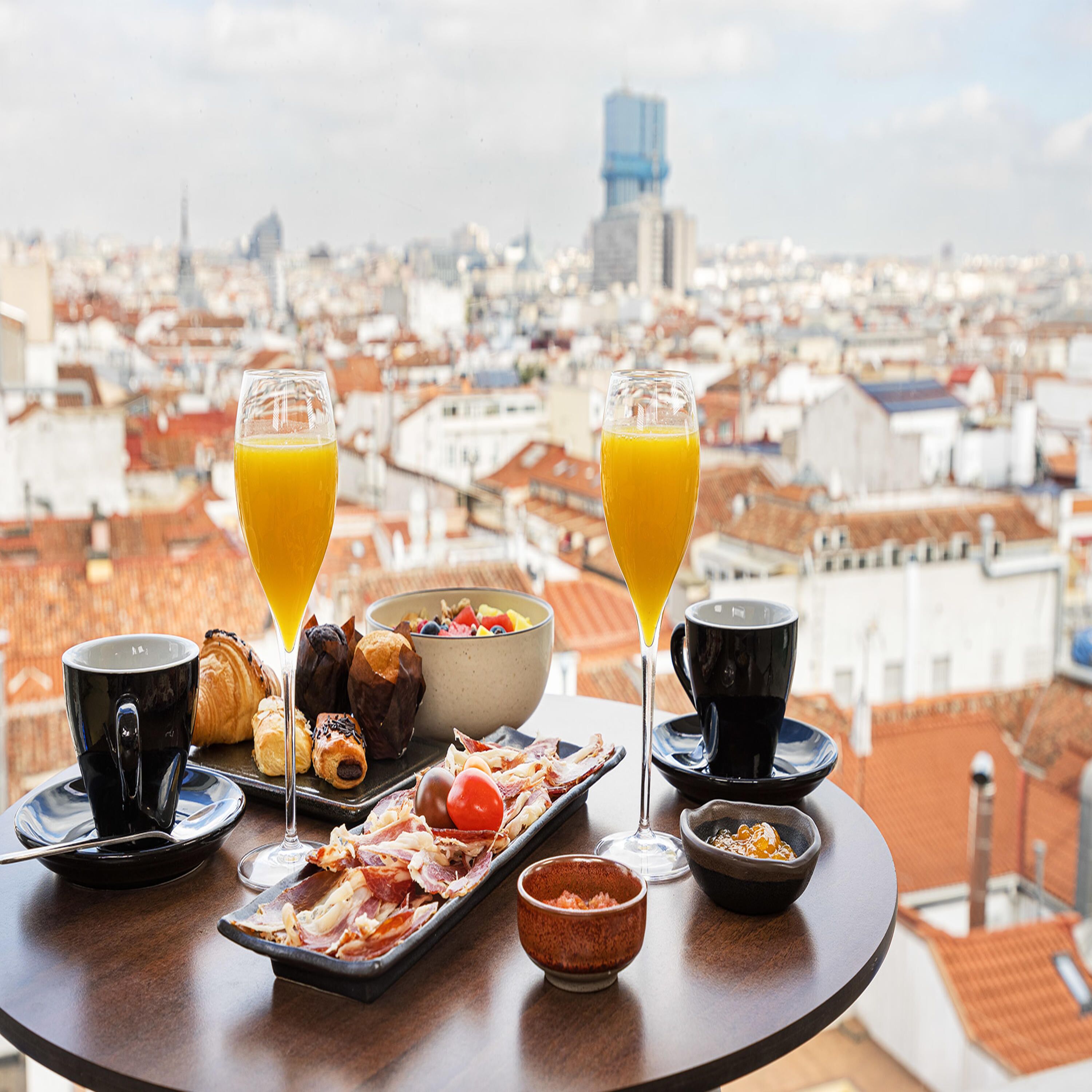A breakfast table with croissants, ham, and a bowl of fruit, overlooking a cityscape.
