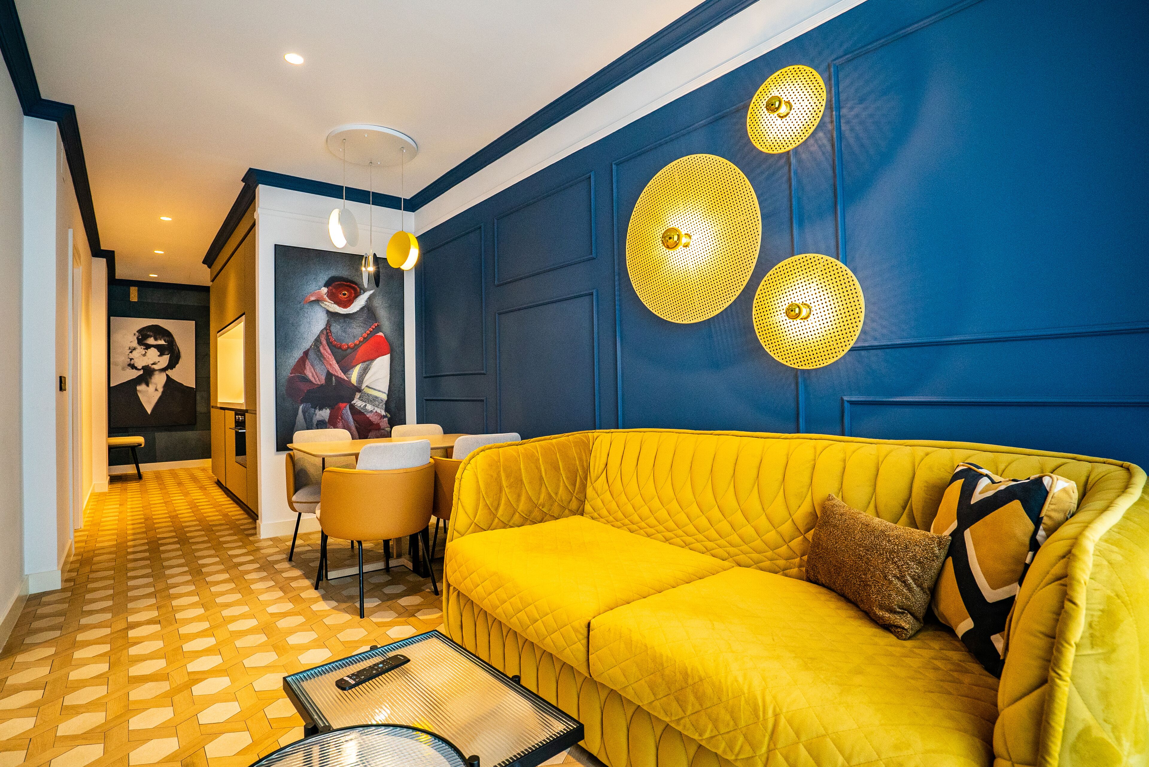A room with a yellow sofa, a dining table, and a painting on the wall.
