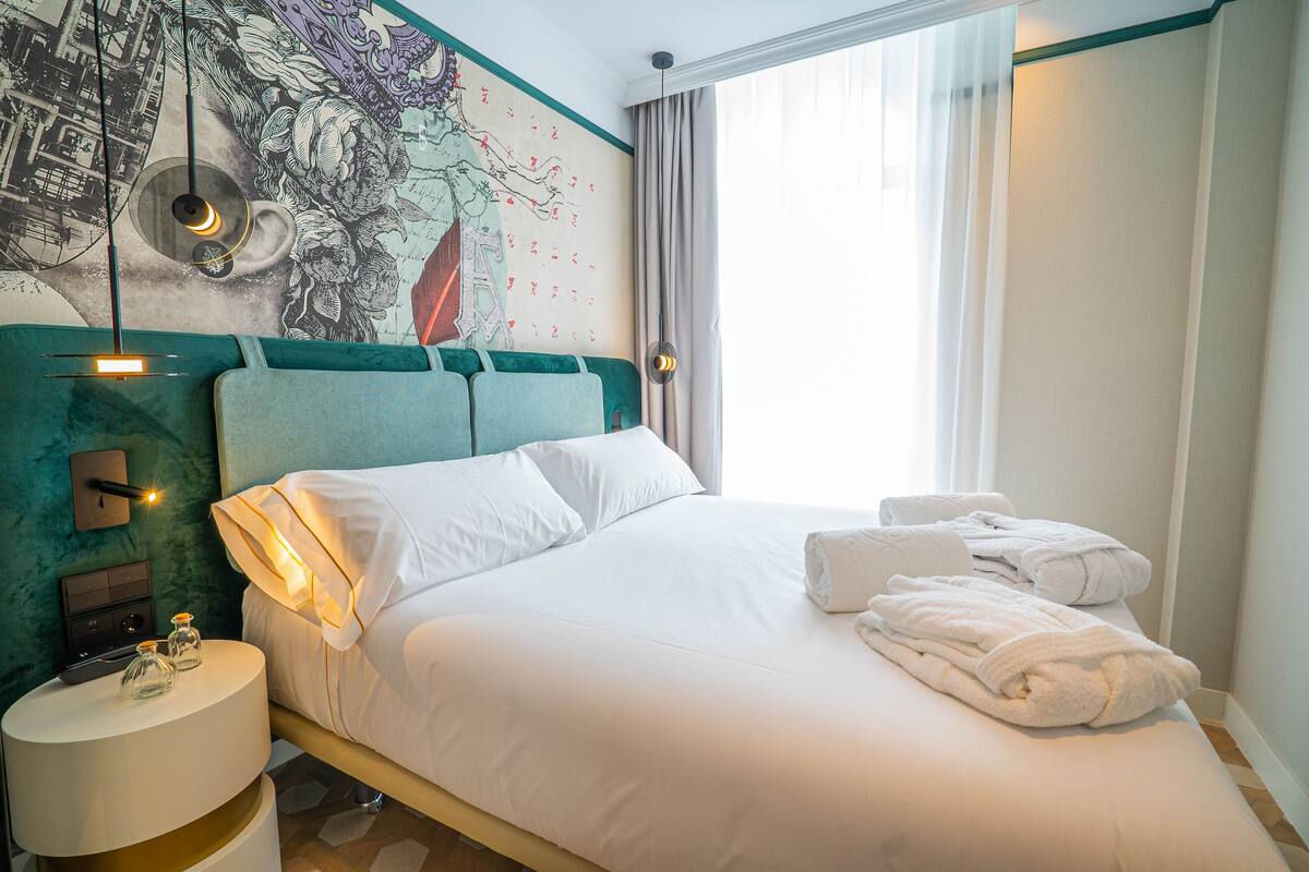 A neatly made bed with white linens and towels, a headboard with a teal accent, a wall-mounted light fixture, and a large window with curtains.