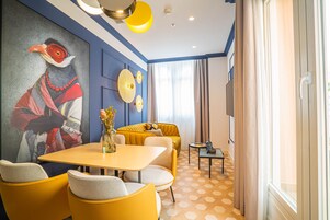 Moderni hotellihuone, jossa on suuri lintukuva, keltainen sohva, ruokapöytä ja kuviollinen lattia.