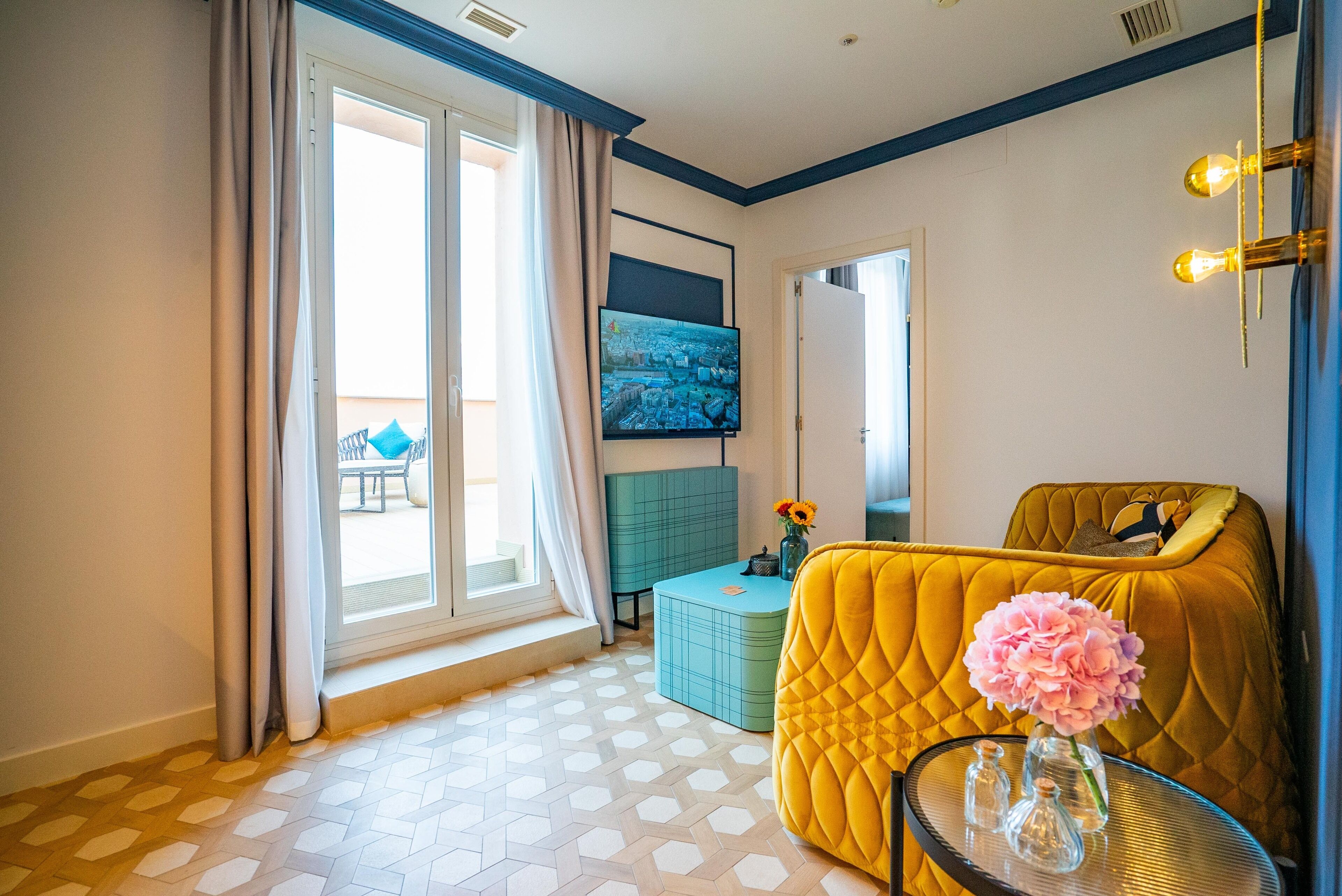 A hotel room with a large window, a television, a yellow armchair, a small table with a vase, and a patterned floor.