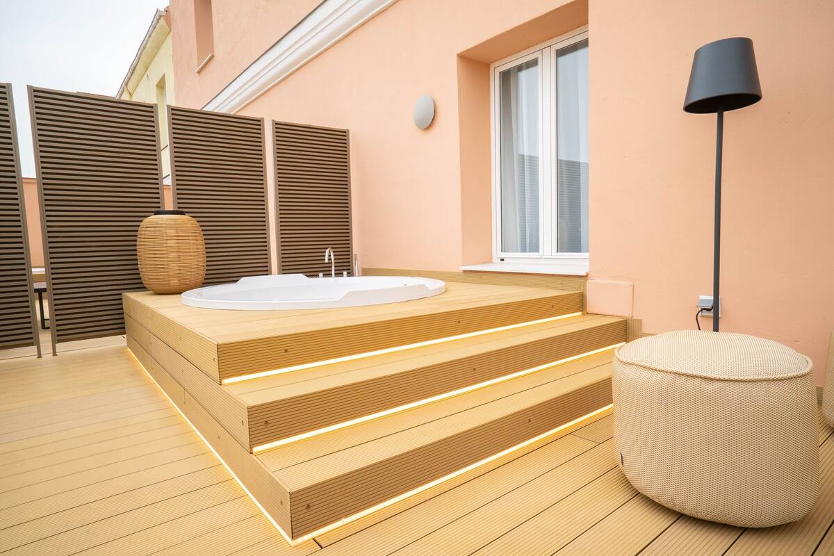 A modern outdoor bathroom with a wooden platform, a hot tub, and a round wicker ottoman.