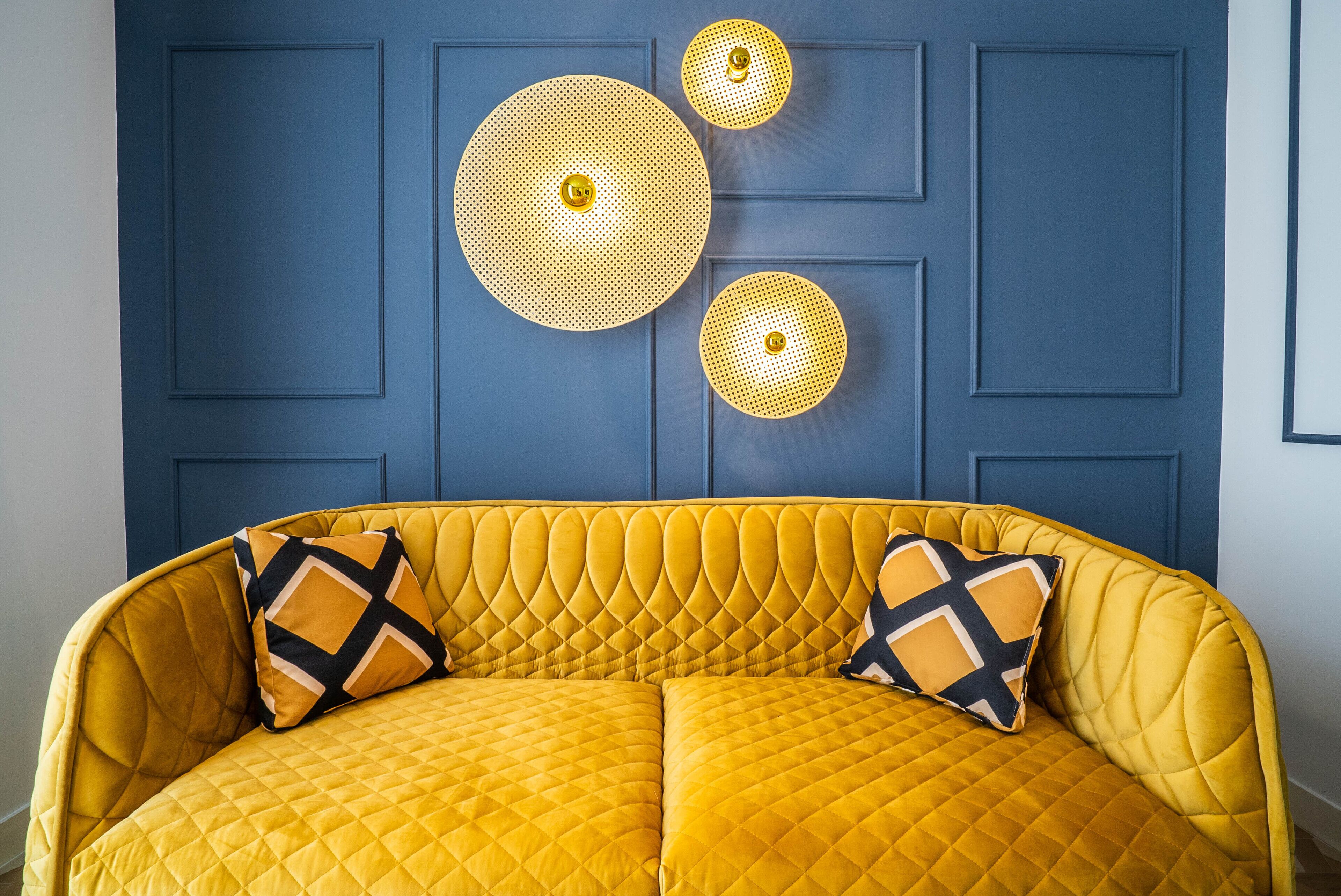 Un sofá amarillo con tapicería acolchada y dos cojines estampados, frente a una pared azul con paneles y tres lámparas esféricas fijadas a la pared.