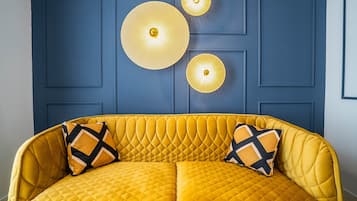 Un sofá amarillo con tapicería acolchada y dos cojines estampados, frente a una pared azul con paneles y tres lámparas esféricas fijadas a la pared.