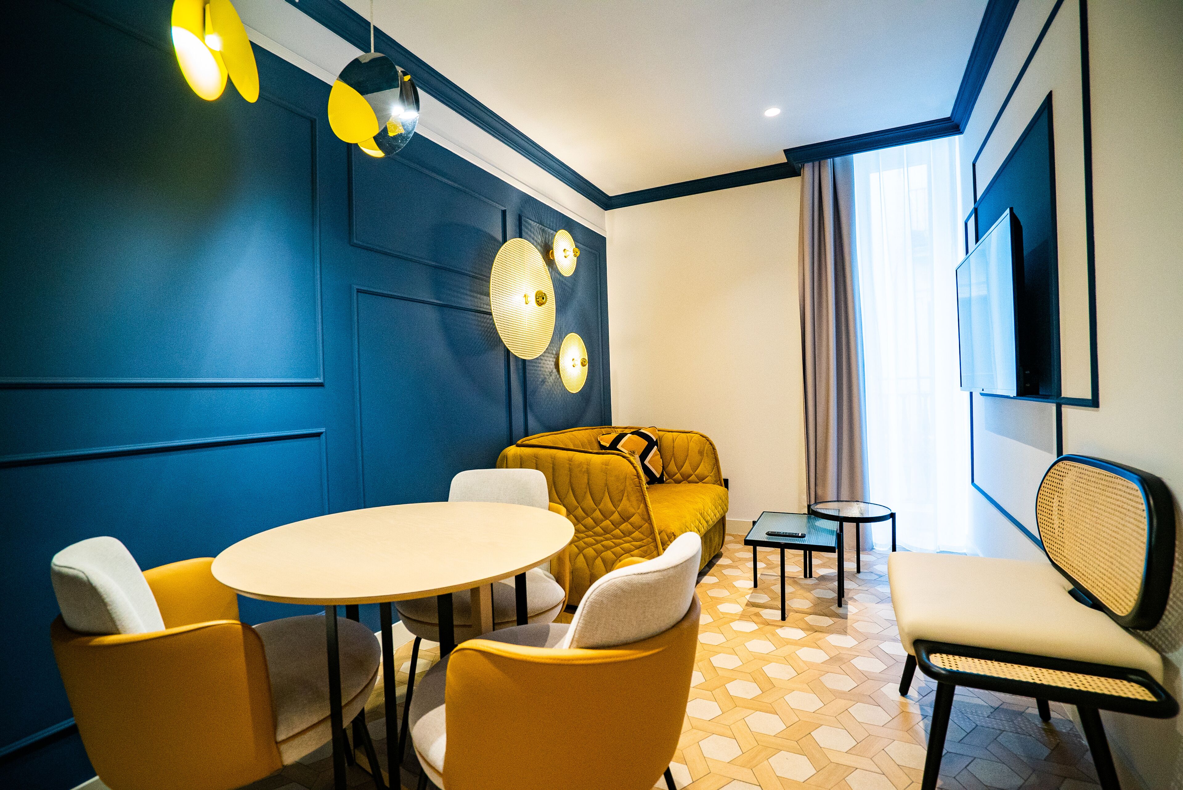 A modern hotel room with a blue accent wall, a round dining table, yellow chairs, a sofa, a small side table, and a flat-screen TV mounted on the wall.