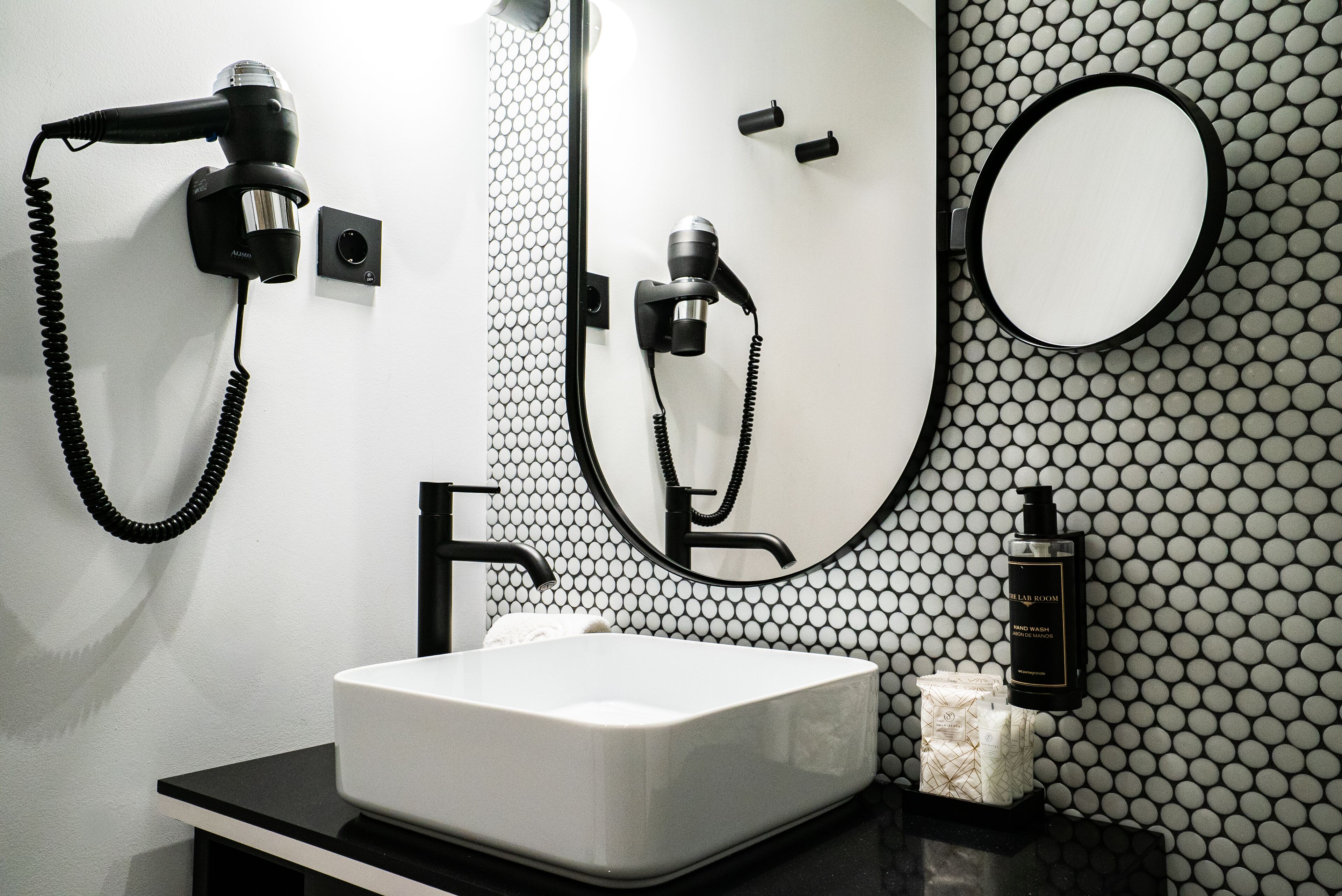 A modern bathroom with a white sink, black countertop, and a mirror with a black frame. There is a hairdryer and a bottle of shampoo on the wall.
