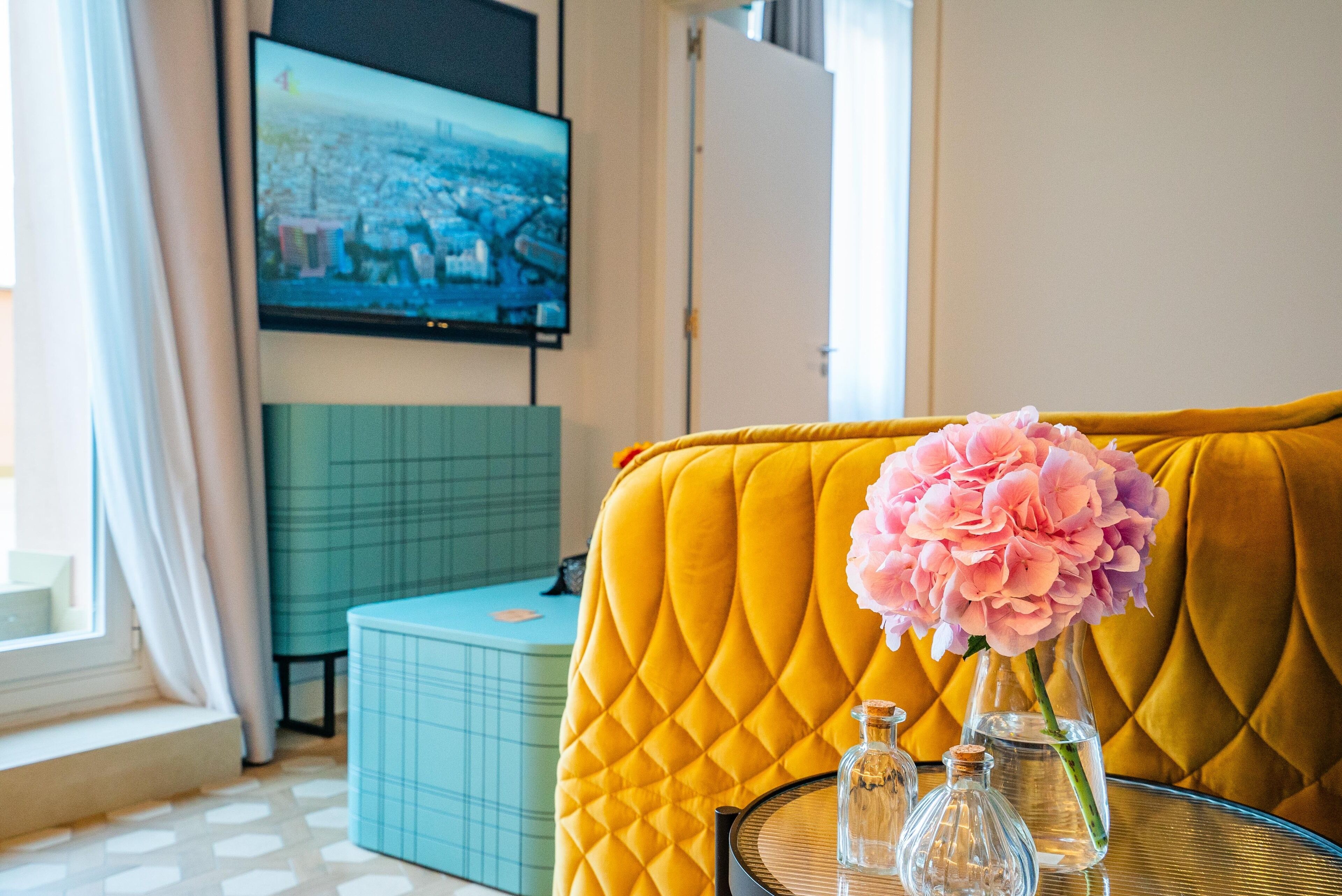 Ein gelbes, gepolstertes Sofa mit einer gesteppten Rückenlehne, eine Glasvase mit rosa Blumen und zwei klare Glasflaschen auf einem runden Tisch.