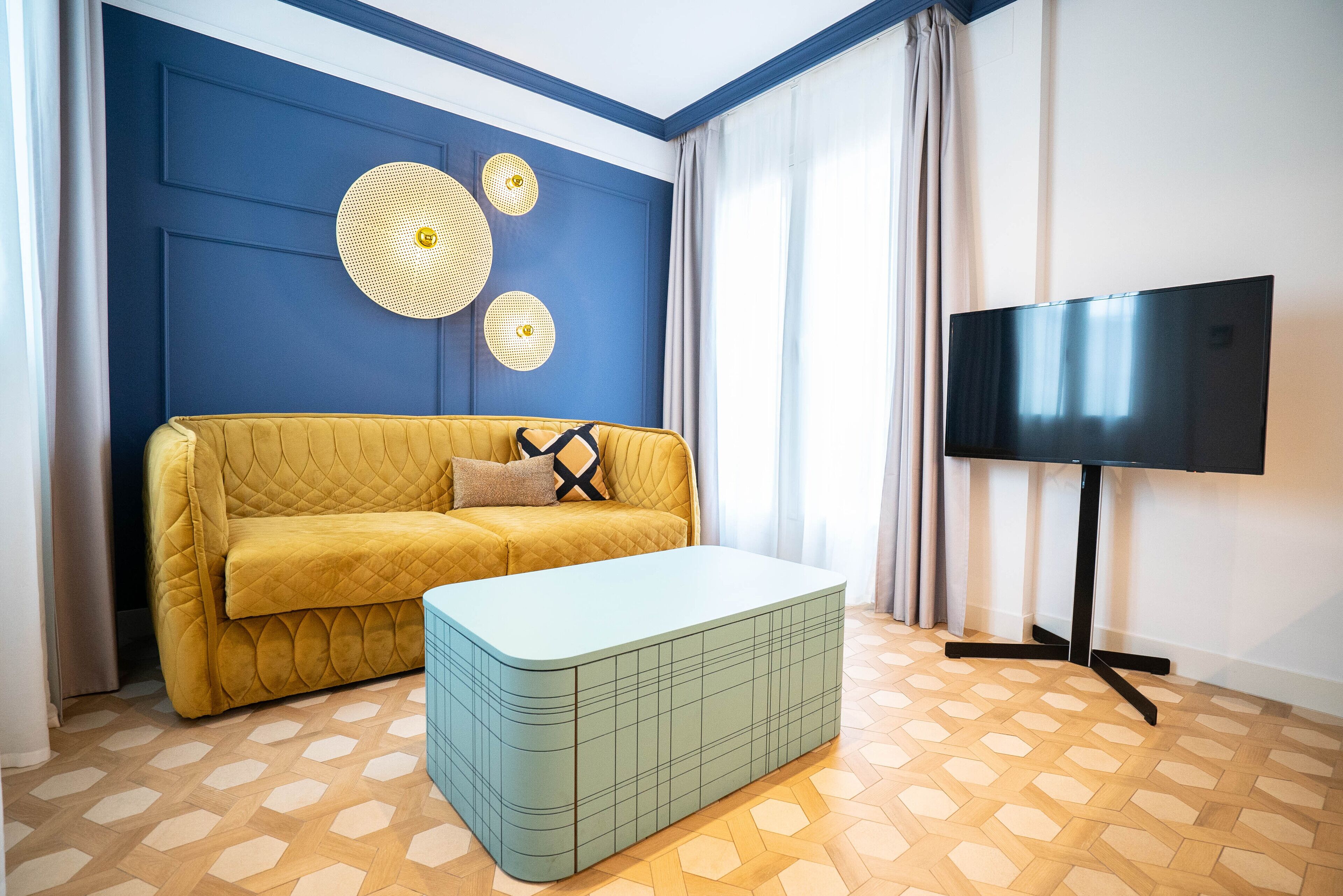 Una stanza con un divano giallo, un pouf azzurro, un televisore a schermo piatto su un supporto e un pavimento di legno decorato.