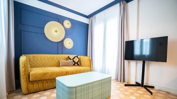 Una stanza con un divano giallo, un pouf azzurro, un televisore a schermo piatto su un supporto e un pavimento di legno decorato.