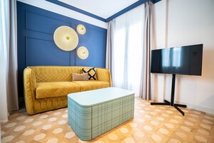 Una stanza con un divano giallo, un pouf azzurro, un televisore a schermo piatto su un supporto e un pavimento di legno decorato.