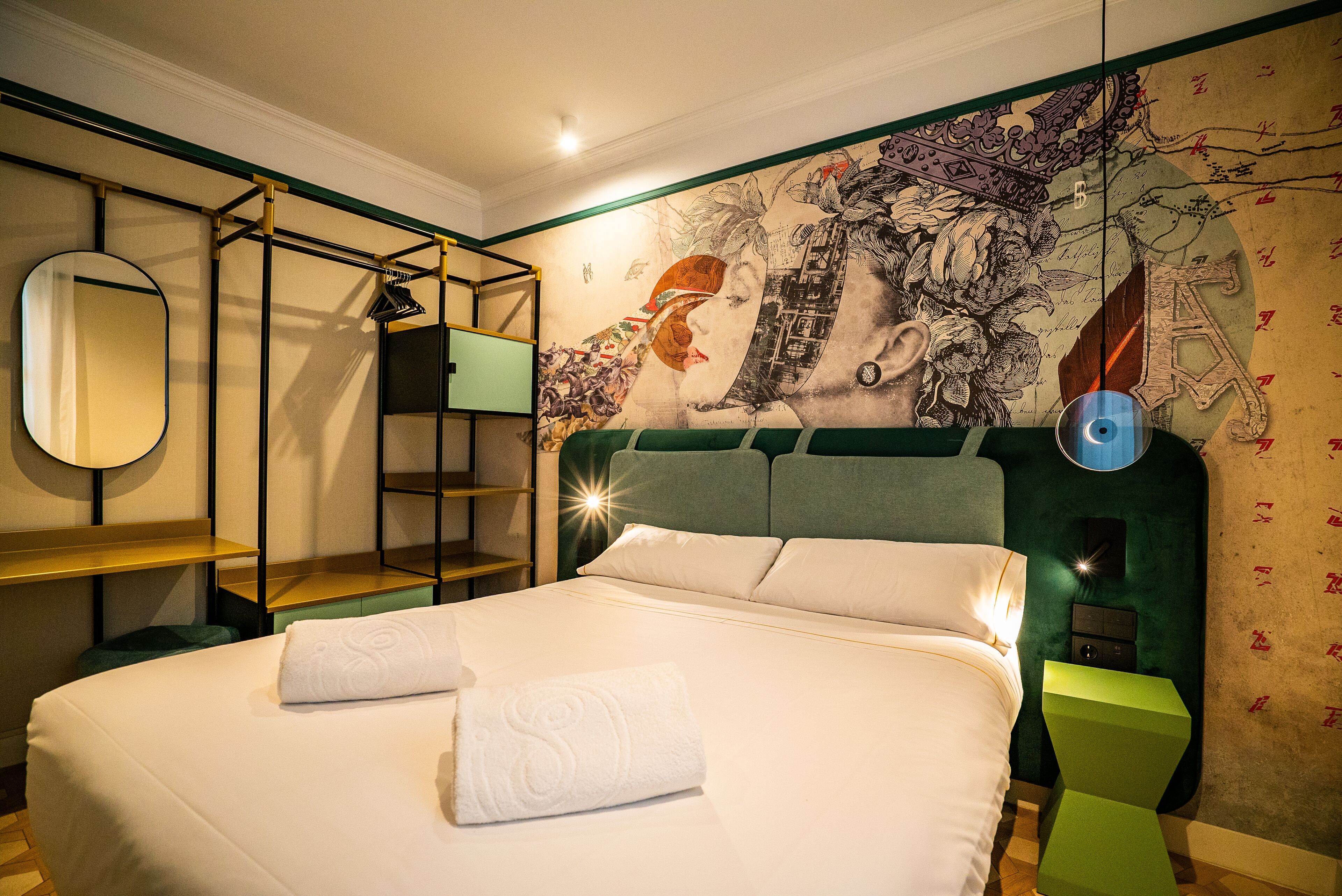 Una cama bien tendida con sábanas blancas y dos toallas, una mesita de noche verde con una lámpara, un estante en la pared y un mural con el retrato de una mujer.