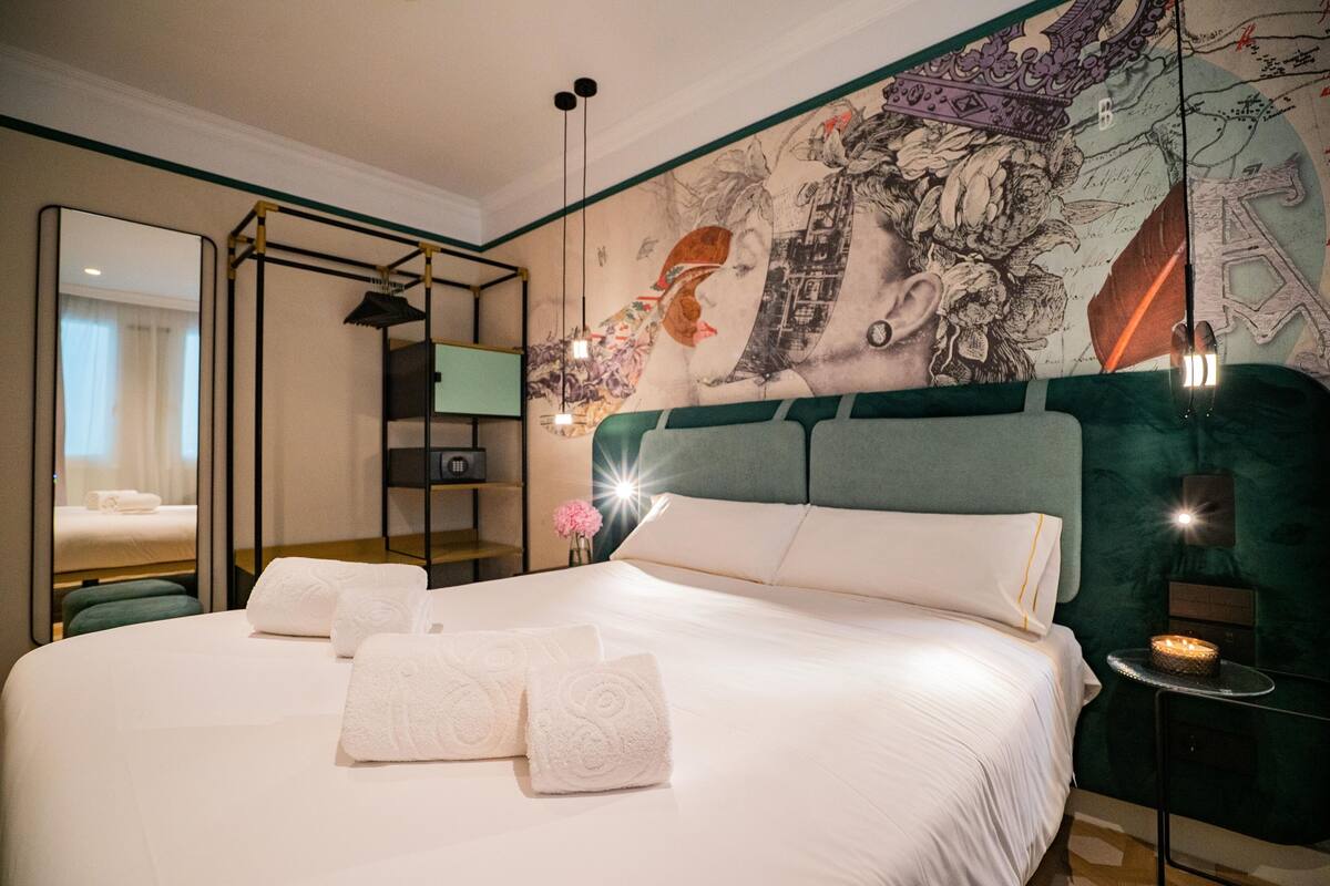 A modern hotel room with a large bed, a mirror, a wall mural, and a small table with a lamp.
