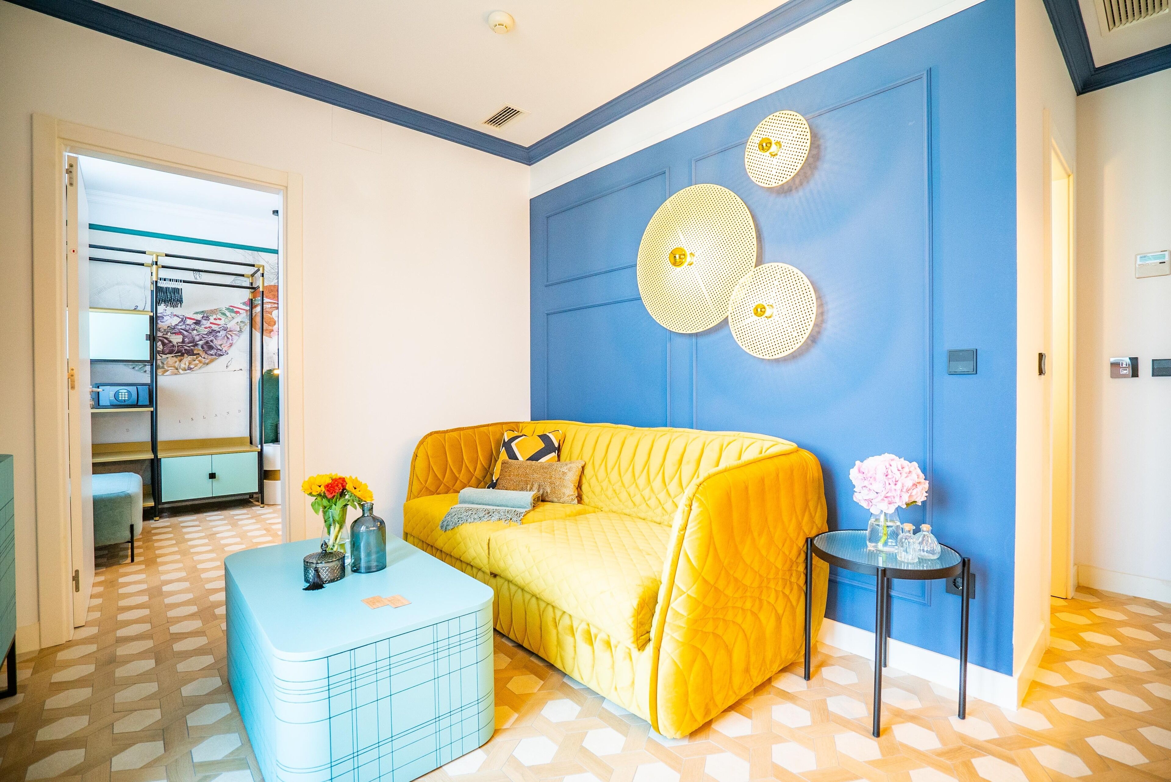Una habitación con un sofá amarillo, un otoman azul y una pequeña mesa con flores. Se aprecia una pared azul con círculos decorativos y un suelo con estampado.
