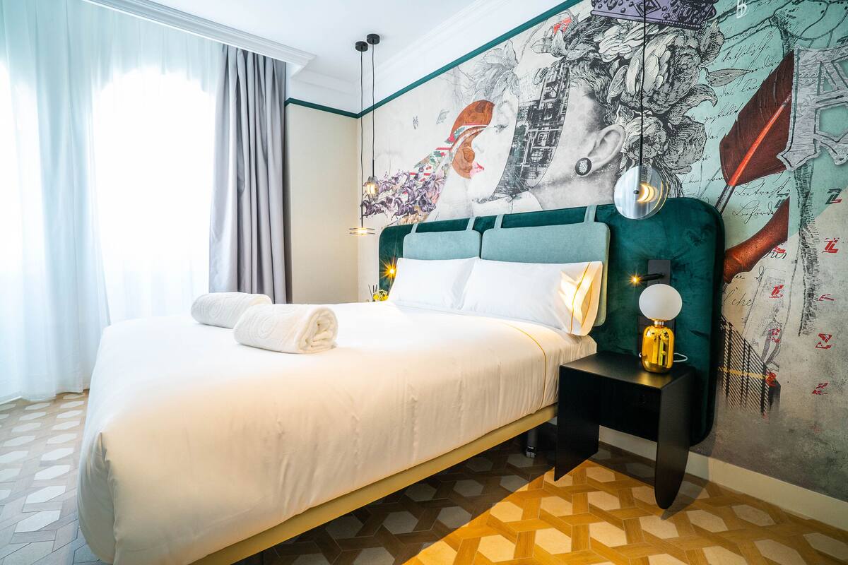 A neatly made bed with white linens and a folded towel, a bedside table with a lamp, and a wall with a mural.