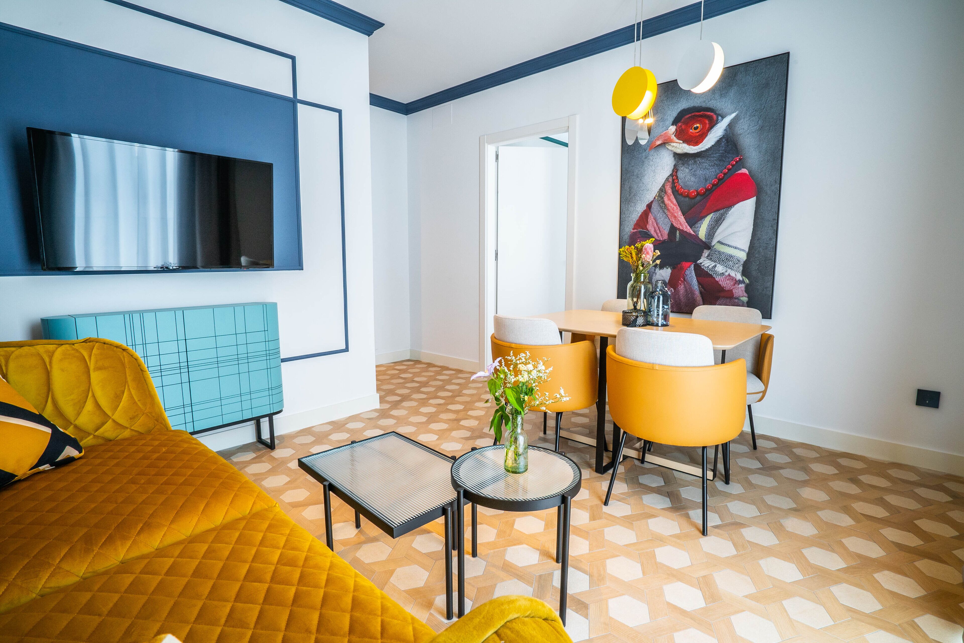 Un moderno salón con un televisor de pantalla plana grande, un sofá amarillo, una pequeña mesa redonda con un jarrón y una zona de comedor con sillas amarillas.