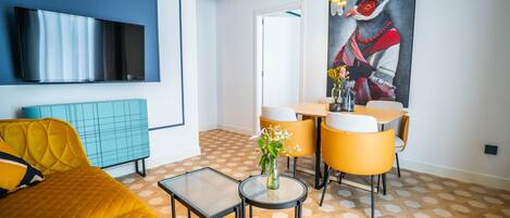Un moderno salón con un televisor de pantalla plana grande, un sofá amarillo, una pequeña mesa redonda con un jarrón y una zona de comedor con sillas amarillas.
