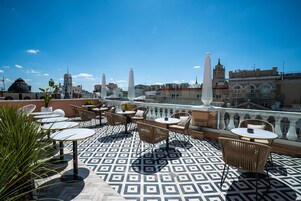 Eine Dachterrasse mit gemustertem Bodenbelag, Sitzgelegenheiten im Freien und Blick auf Stadtgebäude.