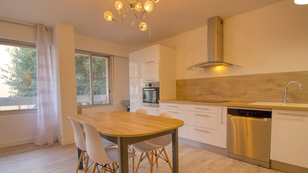 A modern kitchen with a wooden table, white chairs, a stainless steel dishwasher, and a built-in oven.