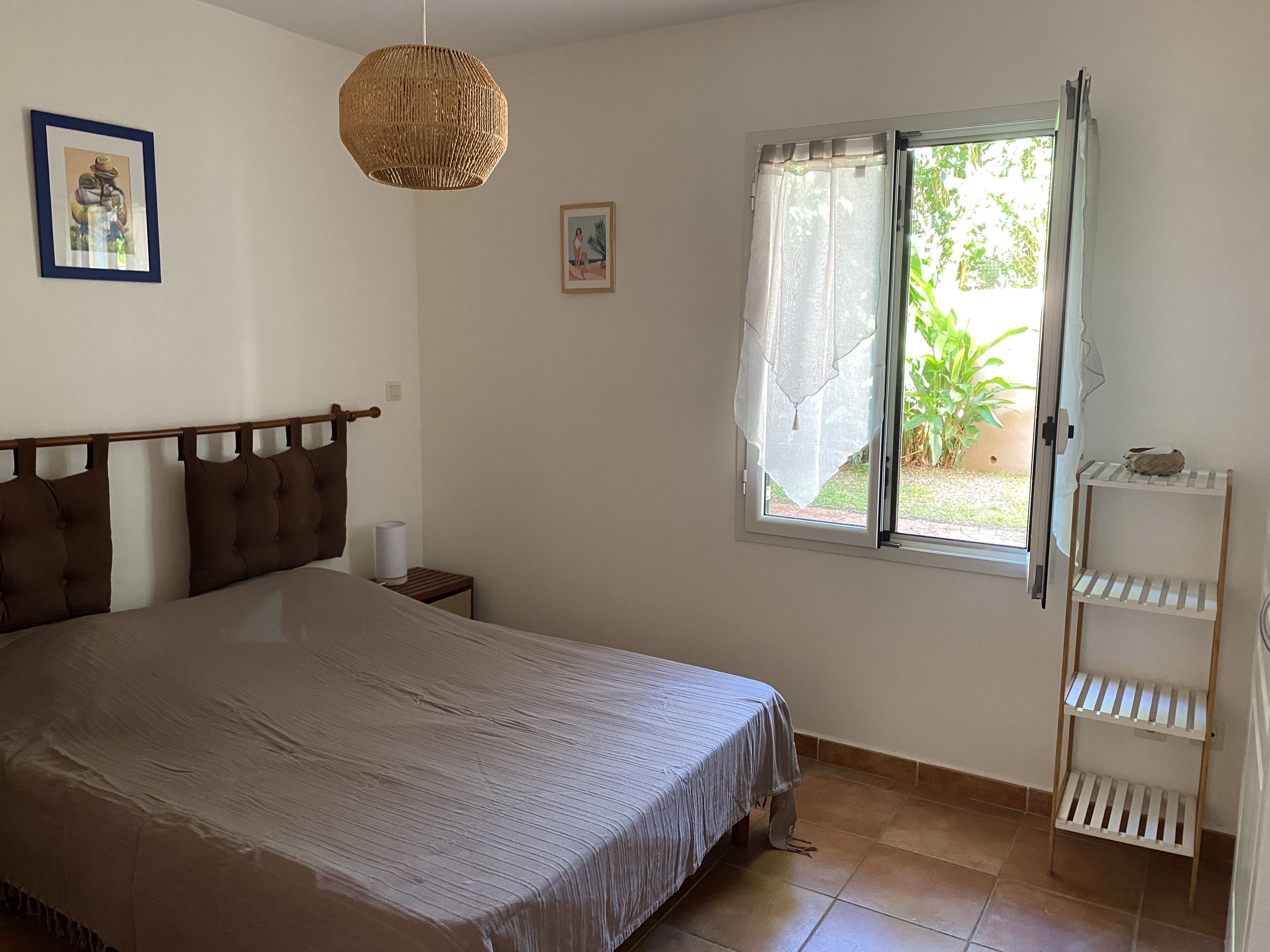 A bedroom with a bed, a bedside table, a window with curtains, a wall-mounted shelf, and a hanging light fixture.