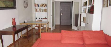 Ein Wohnzimmer mit Holzboden, einem roten Sofa, einem Holztisch, einem Esstisch mit Stühlen, einem Bücherregal und einer Tür zu einem anderen Raum.