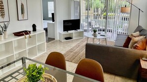 Un salon moderne doté d’un grand luminaire suspendu en fibres tressées, d’un téléviseur à écran plat, d’un canapé et d’un coin repas avec une table et des chaises en verre.