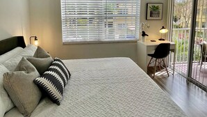 Une chambre à coucher comprenant un lit, un bureau et une vue sur l’extérieur par une fenêtre munie de stores.