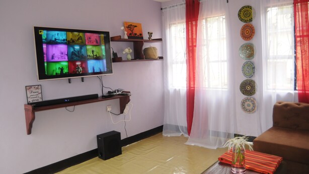 A living room with a flat-screen TV mounted on the wall, wooden shelves with decorative items, a red and white curtain, and a brown sofa. - Hakuna Matata Cozy Apartment (Karatu)
