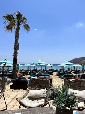 A beachside lounge area with wicker lounge chairs and blue umbrellas, overlooking the sea.