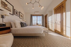 A bedroom with a large bed, a bedside table, a lamp, framed pictures on the wall, and a window with curtains. - Luxury Summer Condo by InvitedHome | Walk to Town, Corner Patio Views (Telluride)