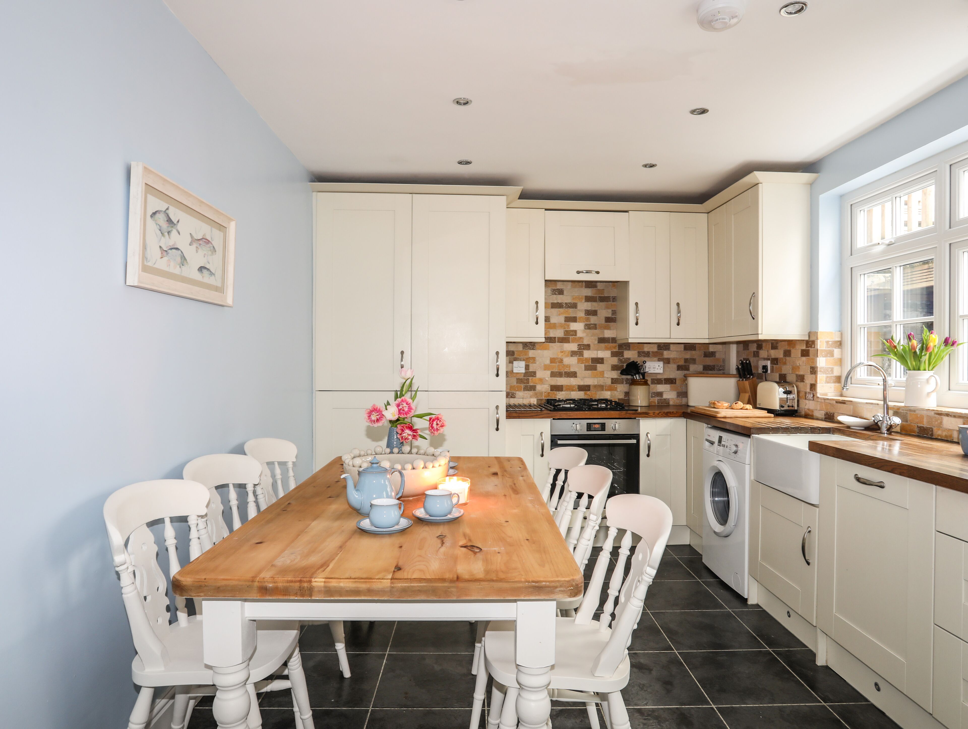 Una cocina con armarios blancos, una mesa de madera y sillas. Hay un fregadero, estufa y lavadora.
