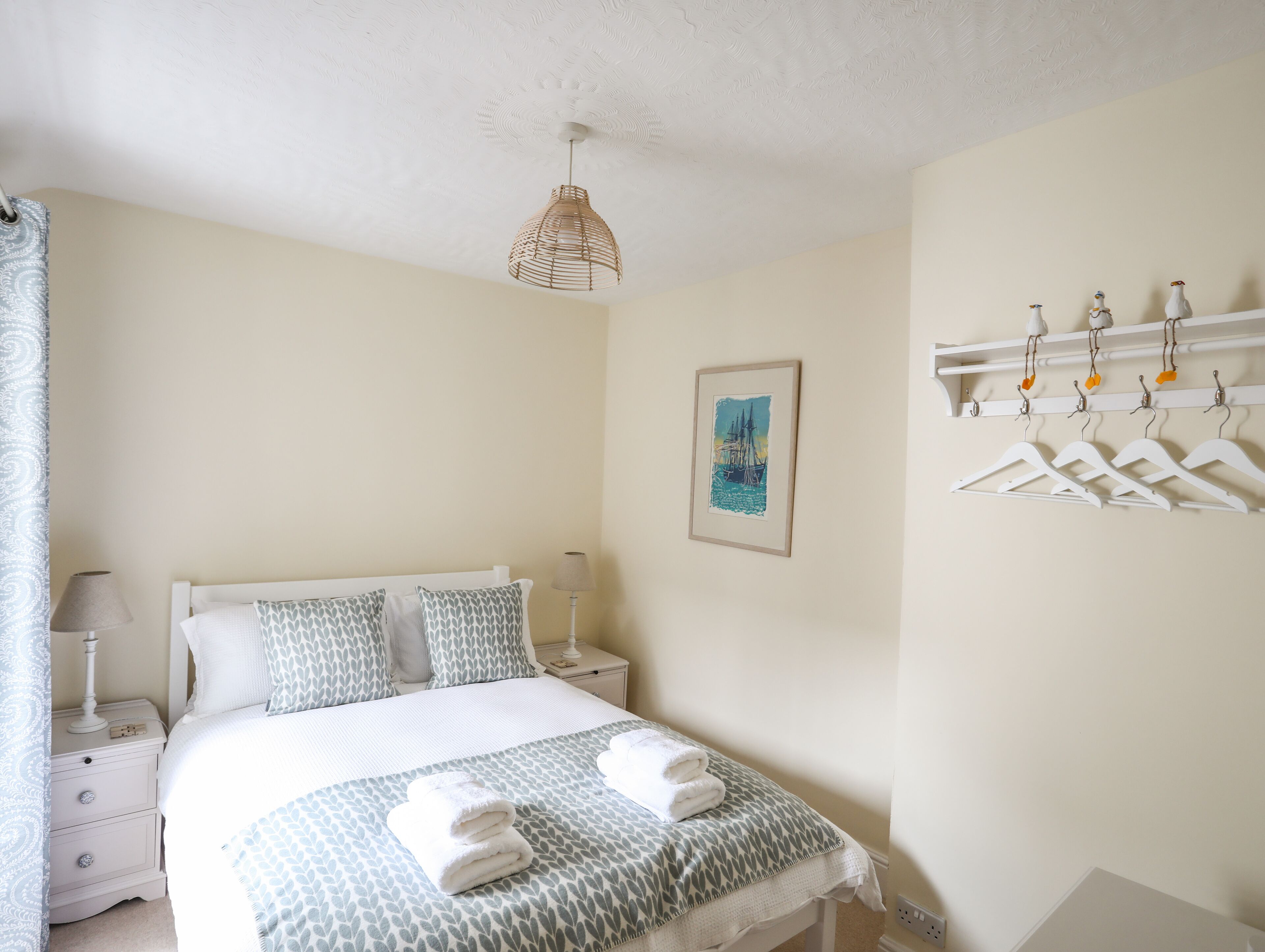 Een netjes opgemaakt slaapkamer met een bed, nachtkastjes, een hanglamp, een kapstok aan de muur en een ingelijst schilderij.